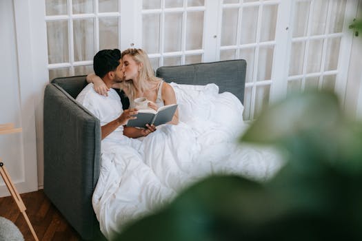 A couple shares an affectionate moment with coffee and a book in a cozy bedroom setting.