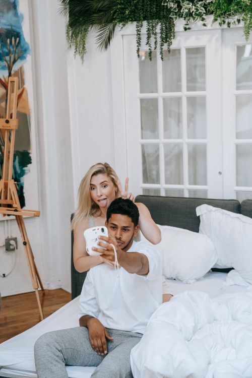 Free Caucasian woman and Hispanic man enjoying a playful moment taking selfies in a bright, cozy bedroom. Stock Photo