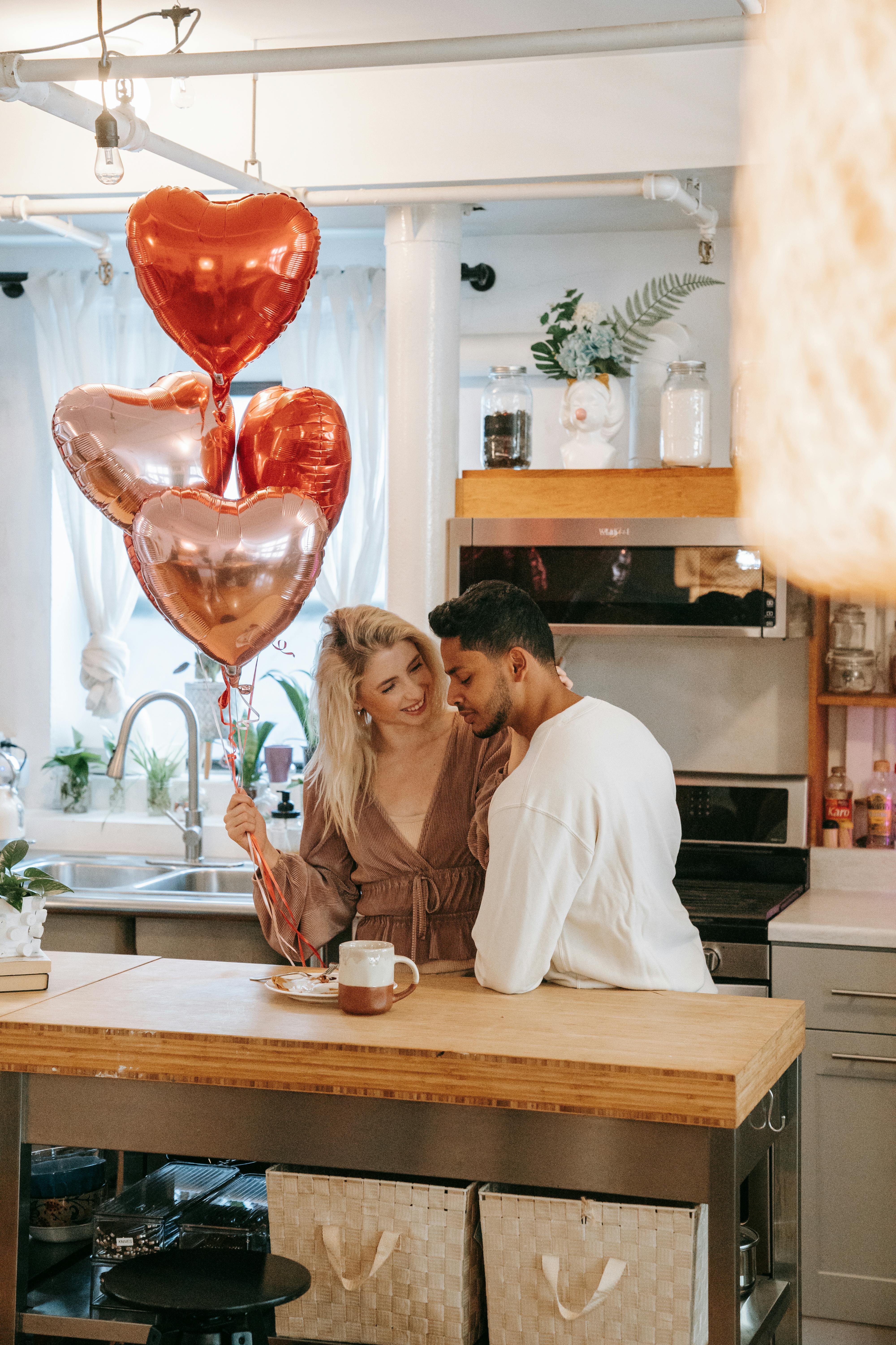 Interracial couple celebrating Valentine's Day in a modern kitchen with heart-shaped balloons.