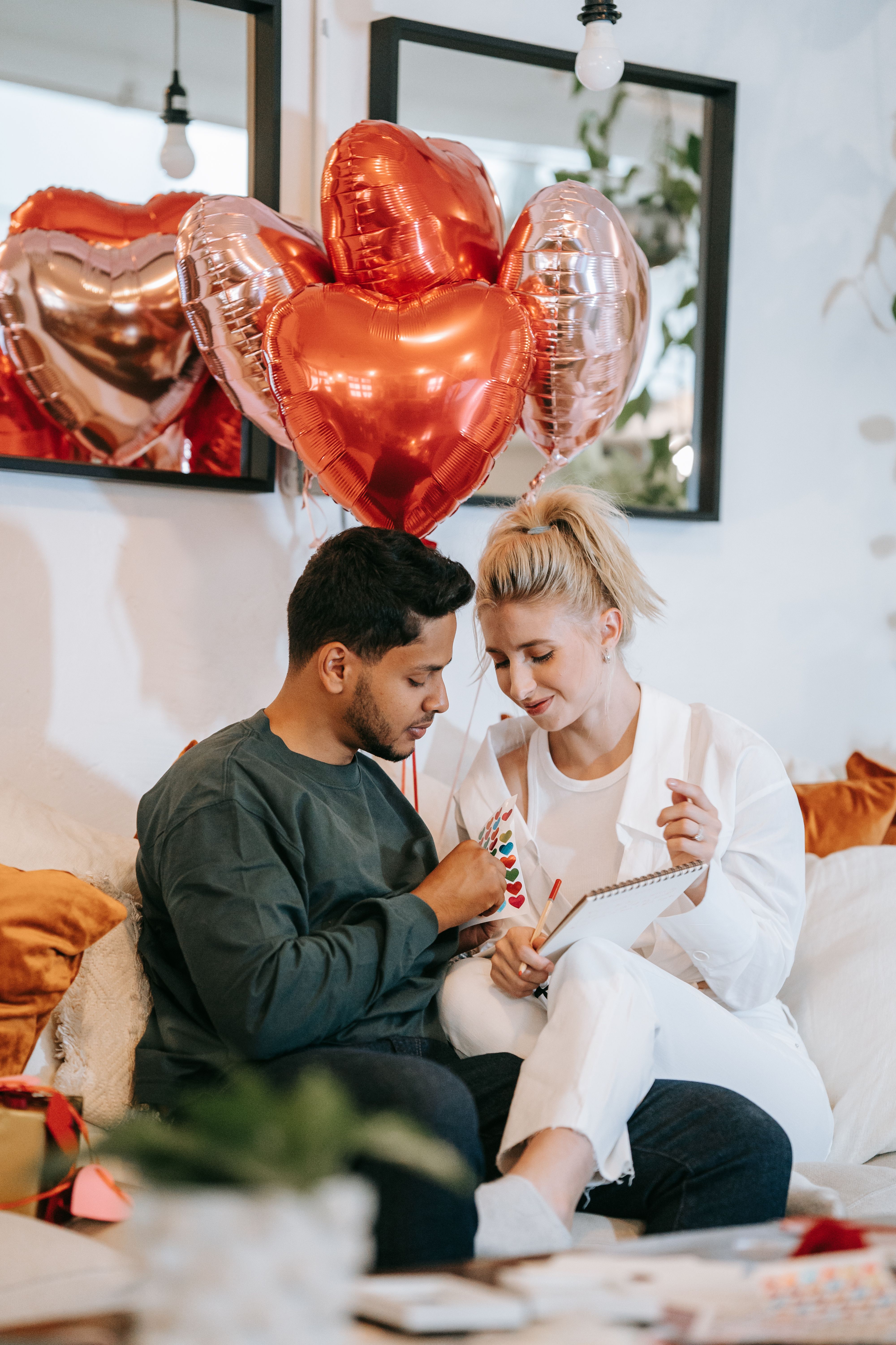 Interracial couple sitting indoors with heart-shaped balloons, enjoying a romantic Valentine's Day moment.
