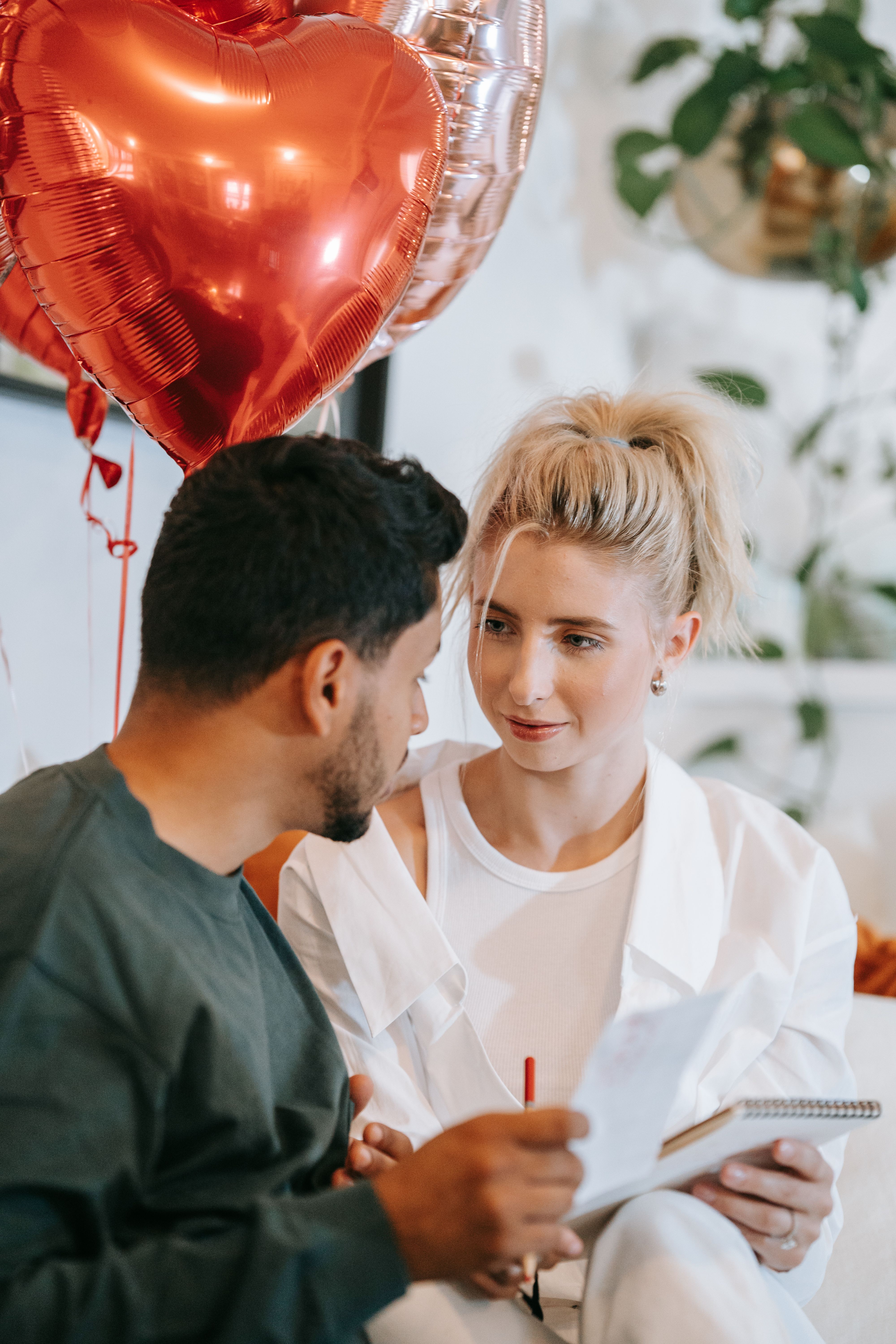 Couple sitting with red heart balloons indoors, sharing a romantic moment.