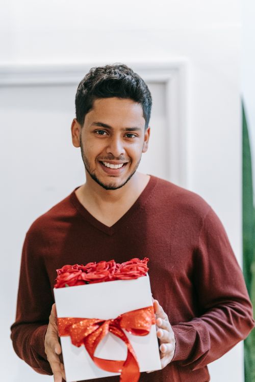 Free Cheerful man with black hair holding a festive gift box indoors. Stock Photo