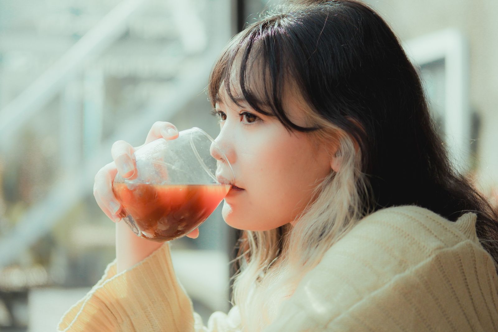 Woman with dark hair and bangs sipping an amber drink from a glass in warm sunlight