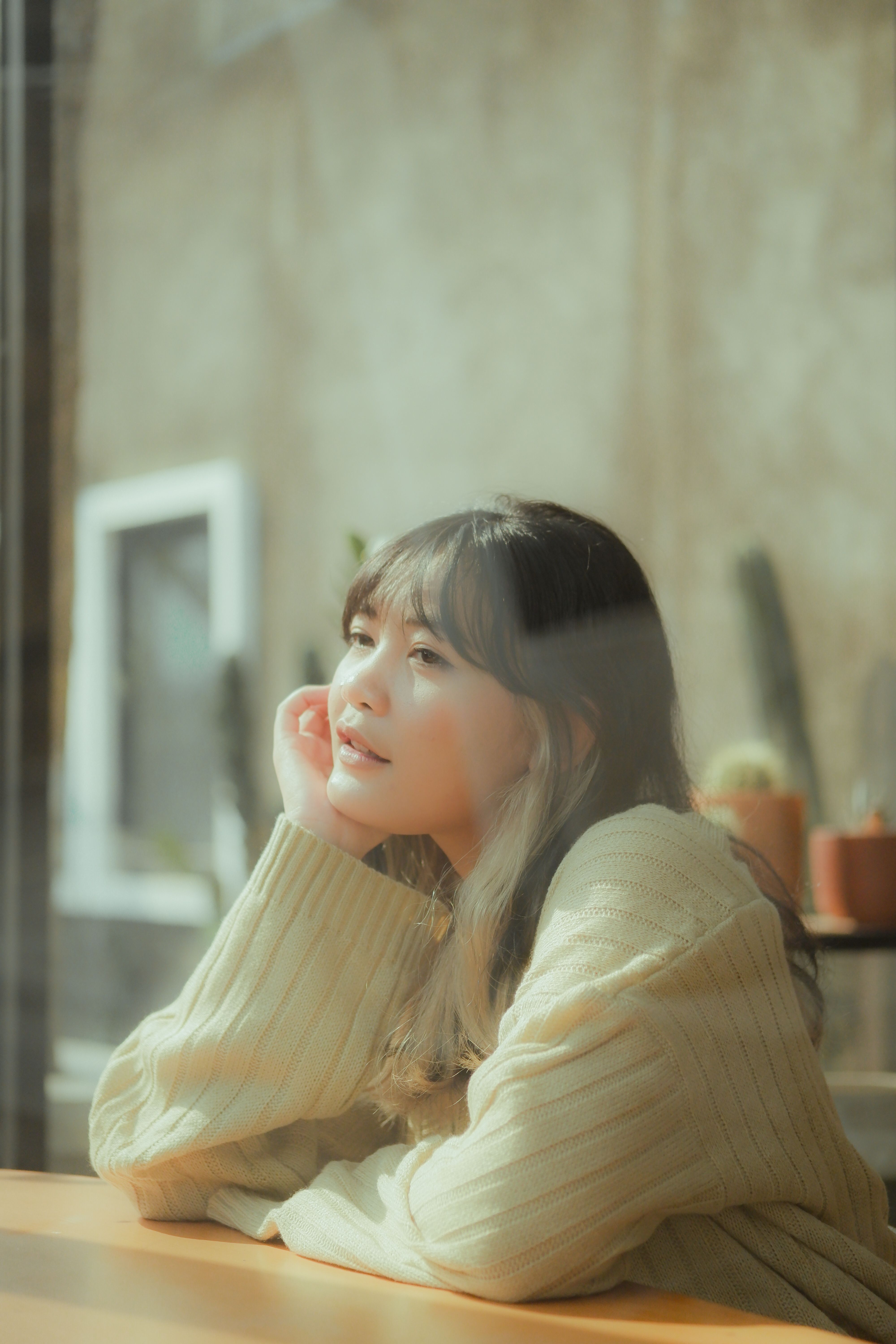 A serene young woman sitting indoors, looking through a sunlit window with a thoughtful expression.