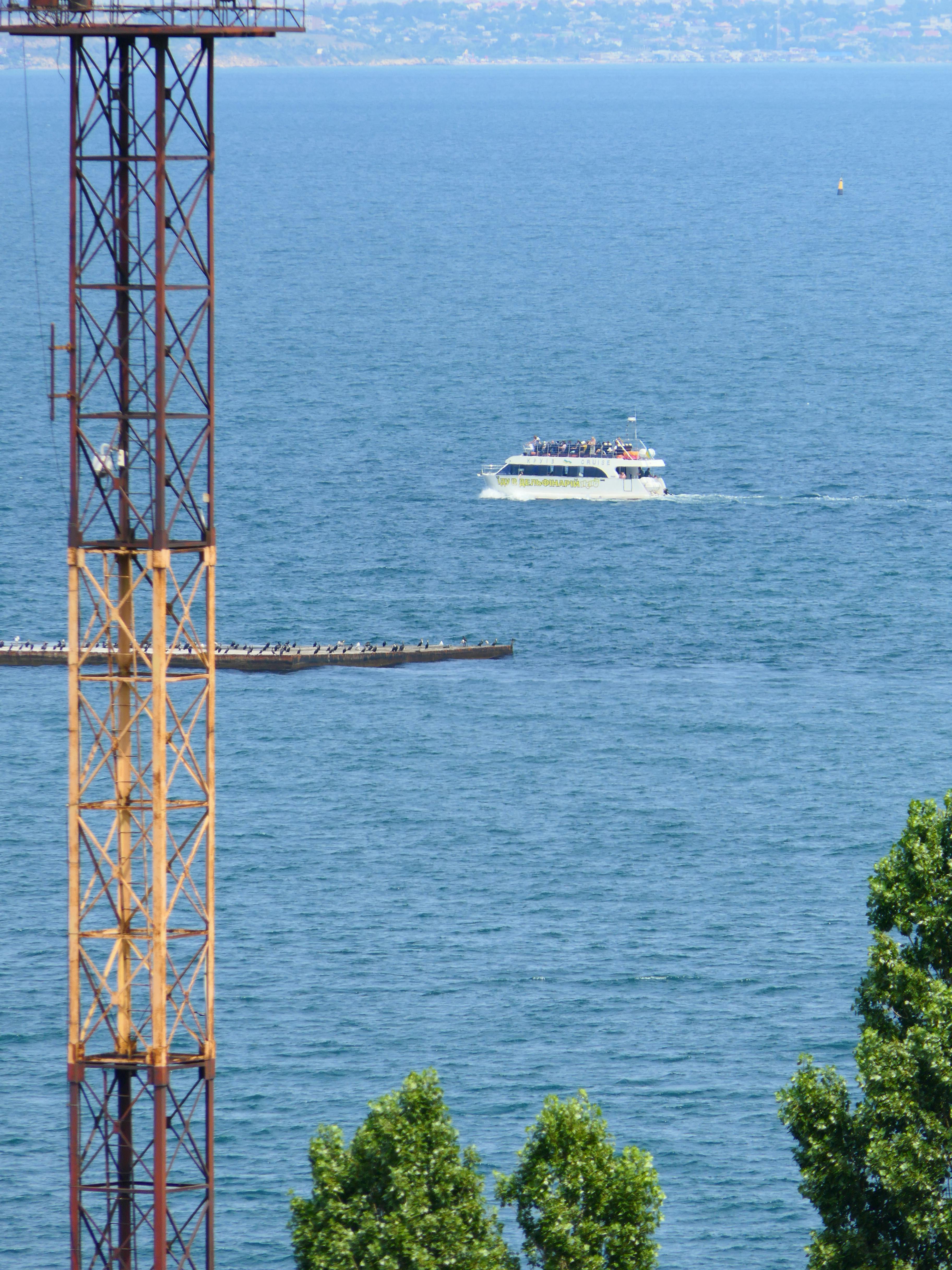 Free A passenger vessel navigating a calm blue ocean with industrial structures and lush greenery in the foreground. Stock Photo