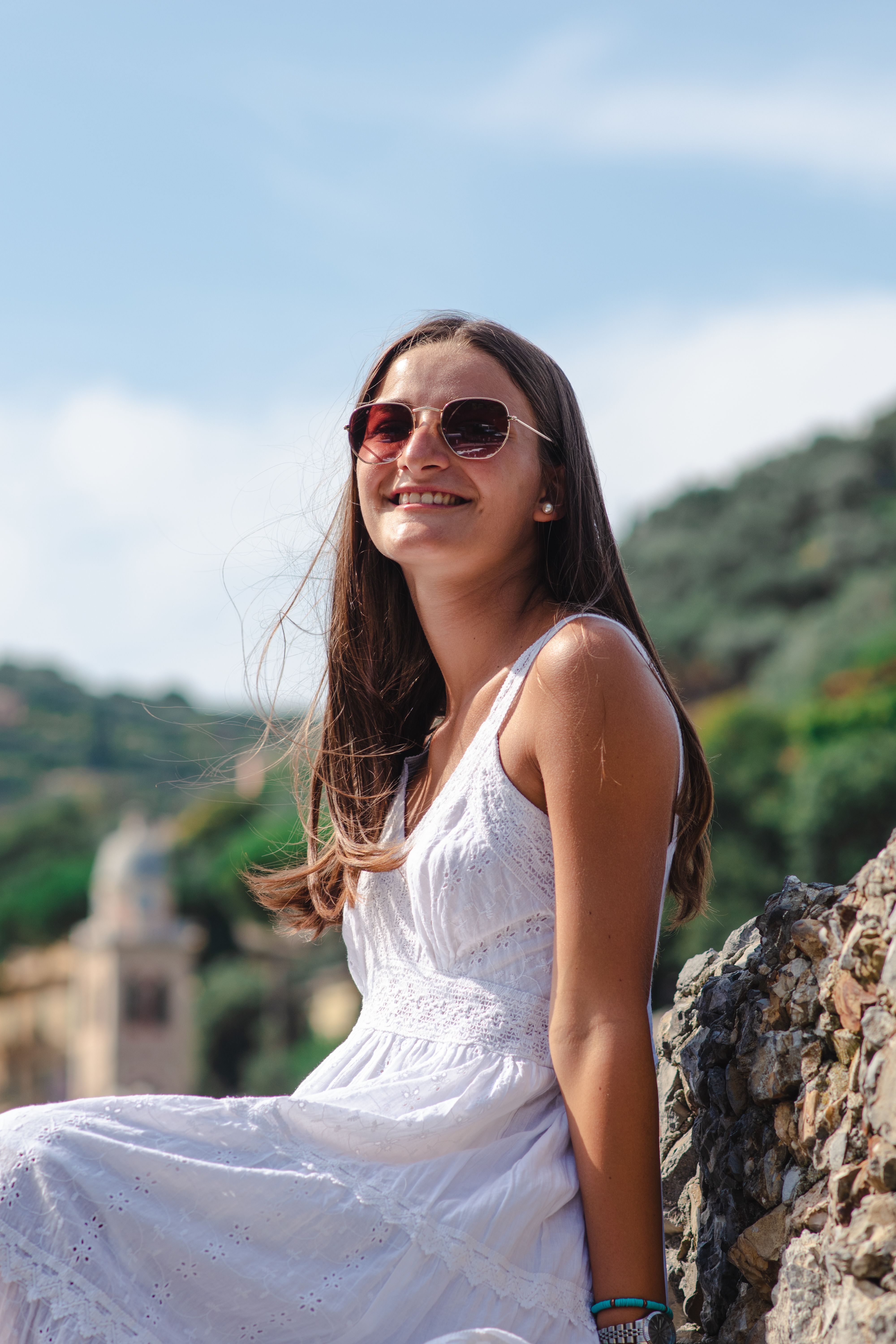 A young woman in a white dress smiles while sitting outdoors on a sunny day.