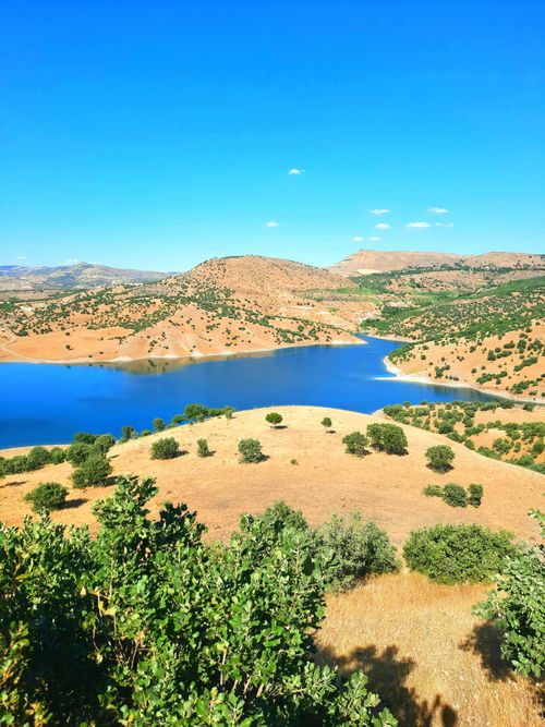 Free A breathtaking aerial view of a tranquil lake surrounded by rolling hills and greenery under a clear blue sky. Stock Photo