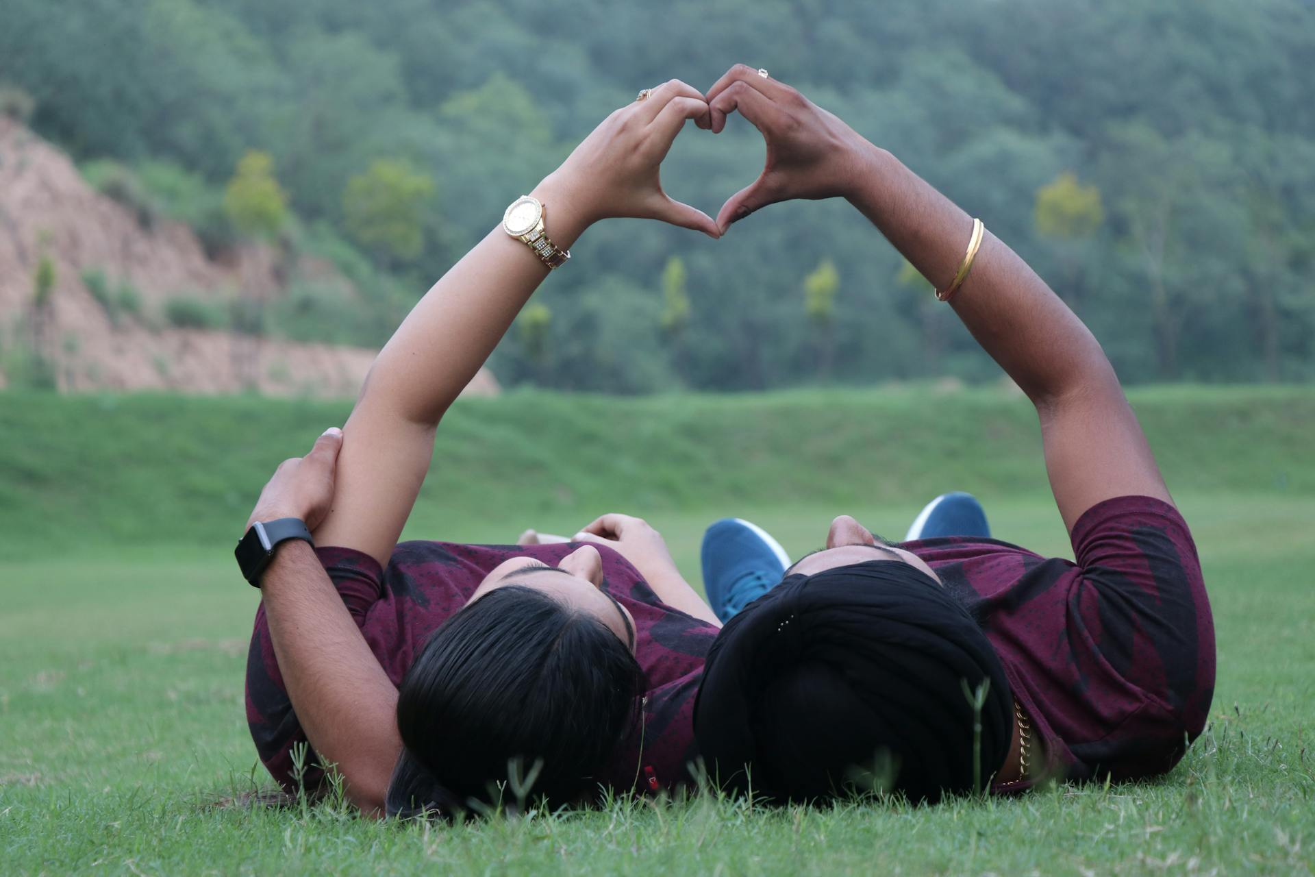 A couple lying in a grassy field forming a heart shape with their hands.