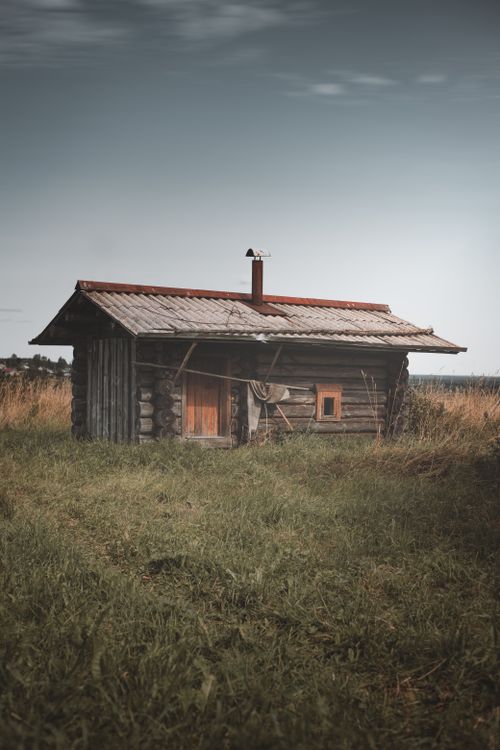 Free A rustic wooden cabin stands alone in a grassy field under a cloudy sky in Russian countryside. Stock Photo