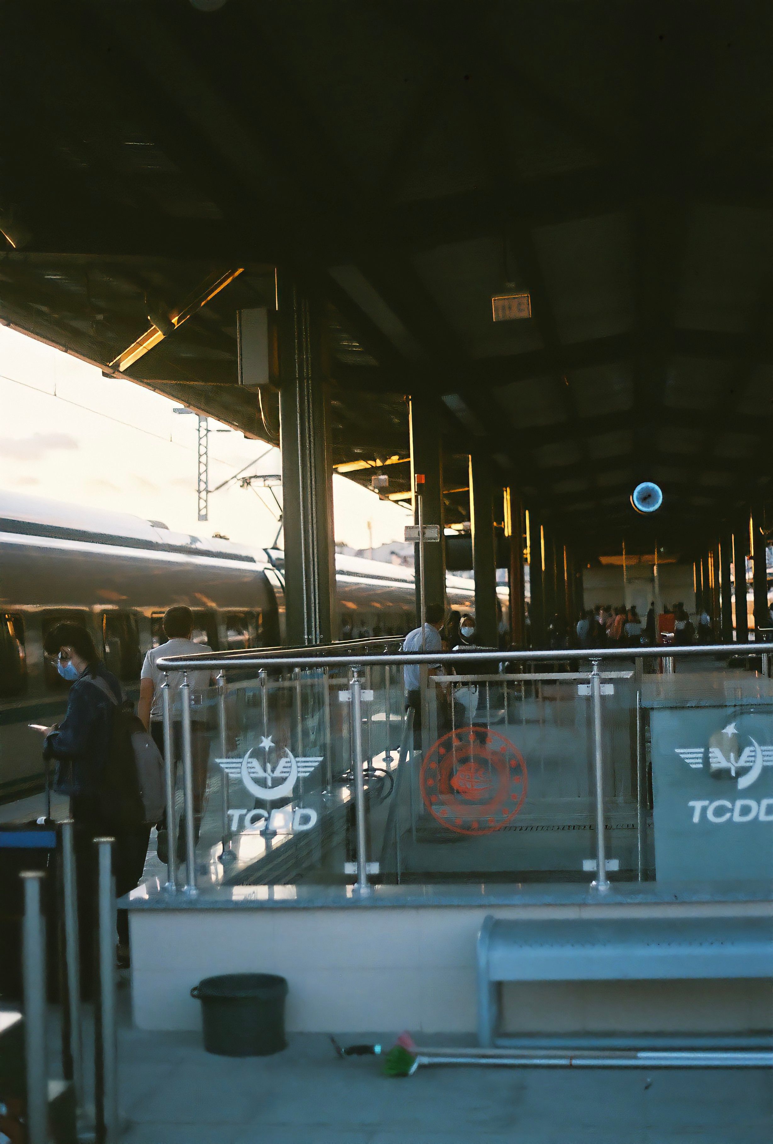A bustling train station with people and a train, under a sunlit platform roof with TCDD branding.