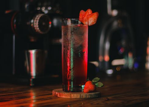 Close-up of a red strawberry cocktail in a glass with ice and fruits on a wooden coaster.
