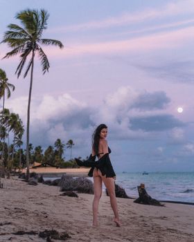Woman enjoying a serene evening on a tropical beach with palm trees, ocean, and full moon in Brazil.