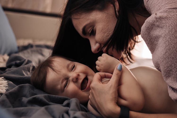 Mother Cuddling A Smiling Baby Lying On A Bed