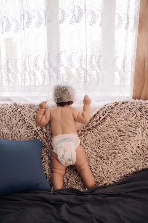 Free Adorable baby in a diaper standing on a couch, curiously peering out a window with sheer curtains. Stock Photo