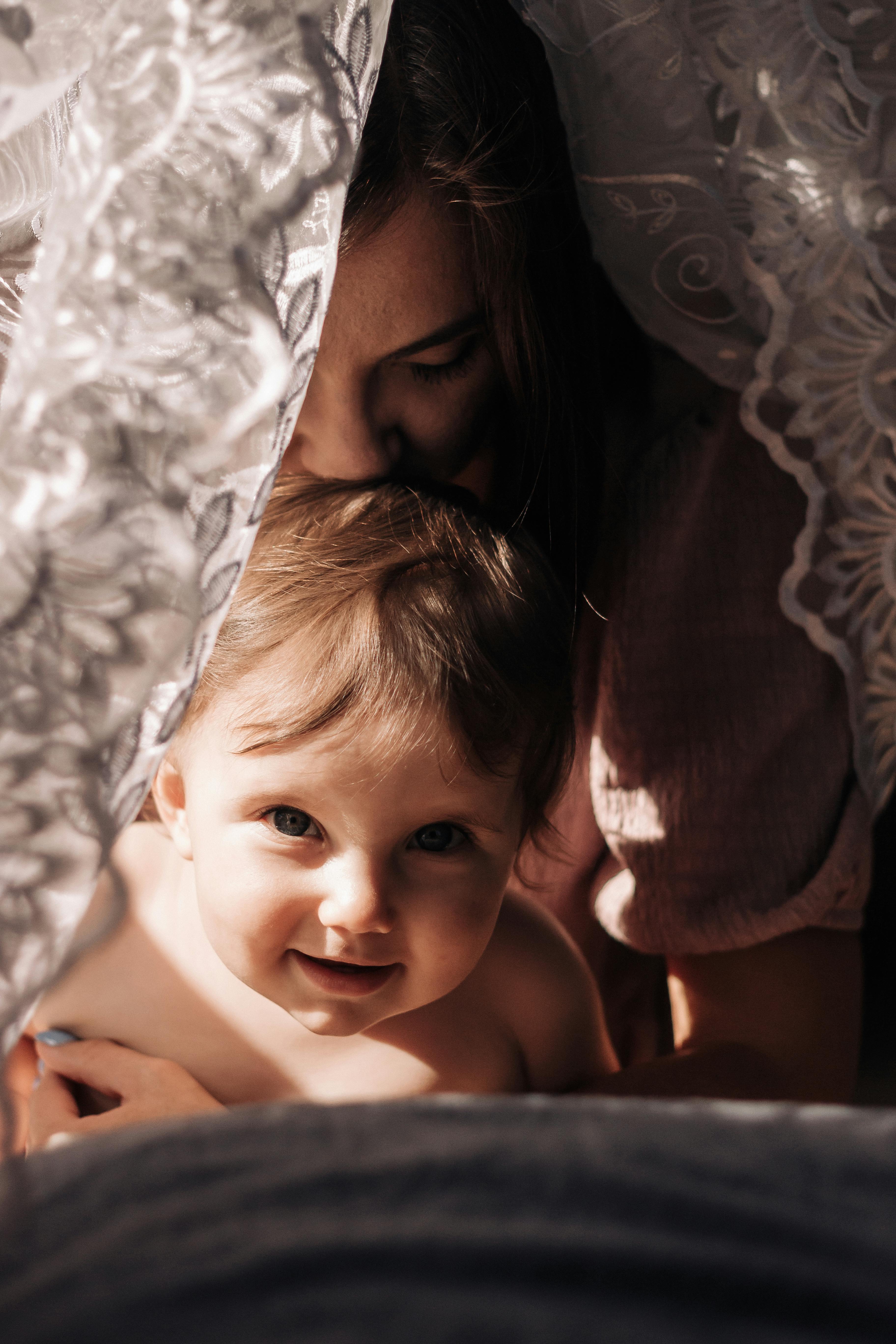 A heartfelt moment of a mother lovingly embracing her baby under a sunlit curtain.