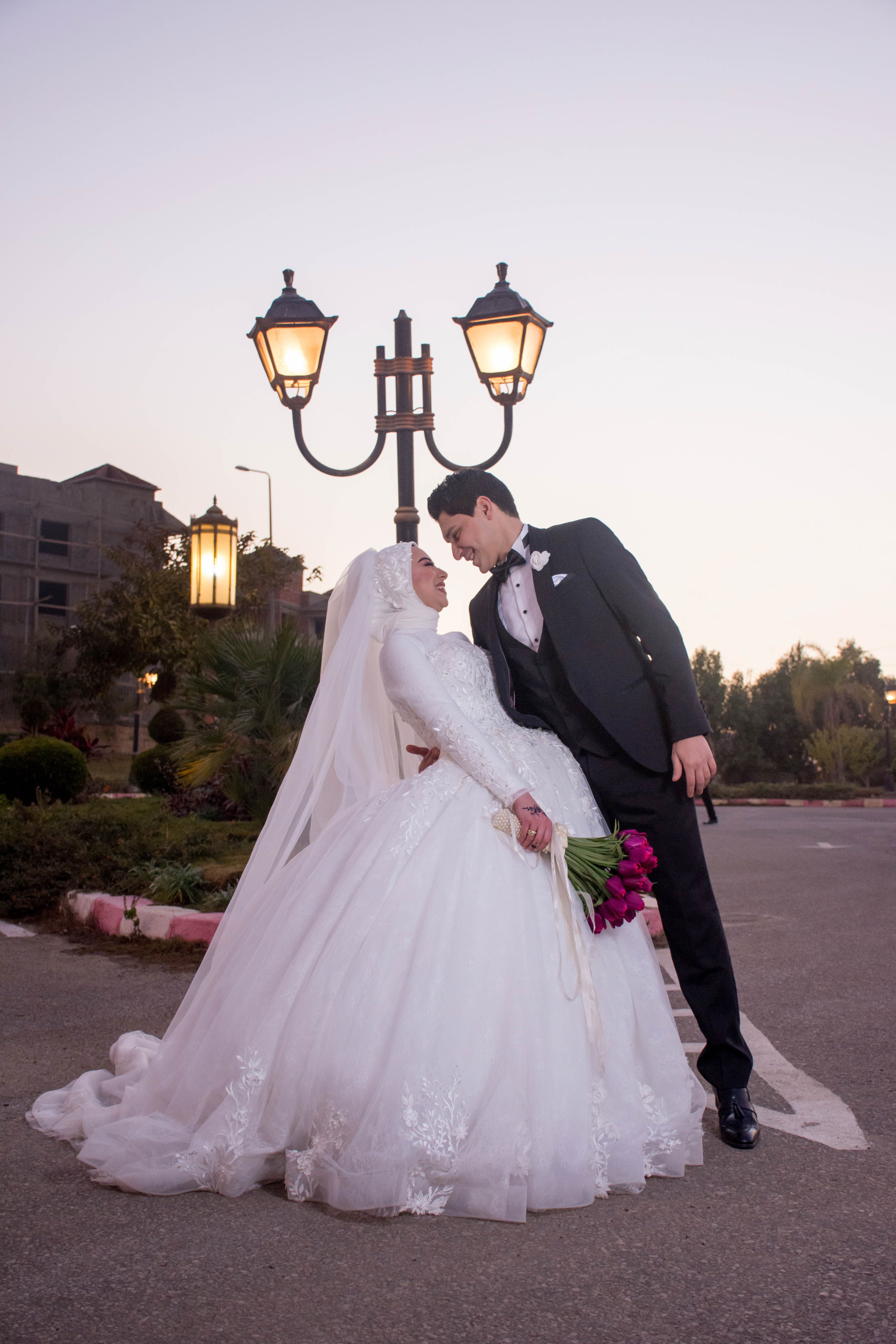 A Muslim bride and groom pose romantically outdoors with street lamps.