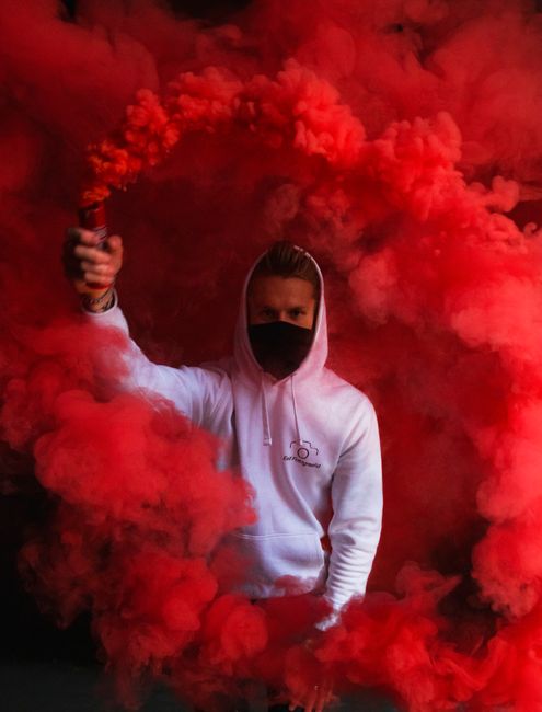 A man in a hoodie and mask holds a red smoke bomb, creating dramatic red clouds.