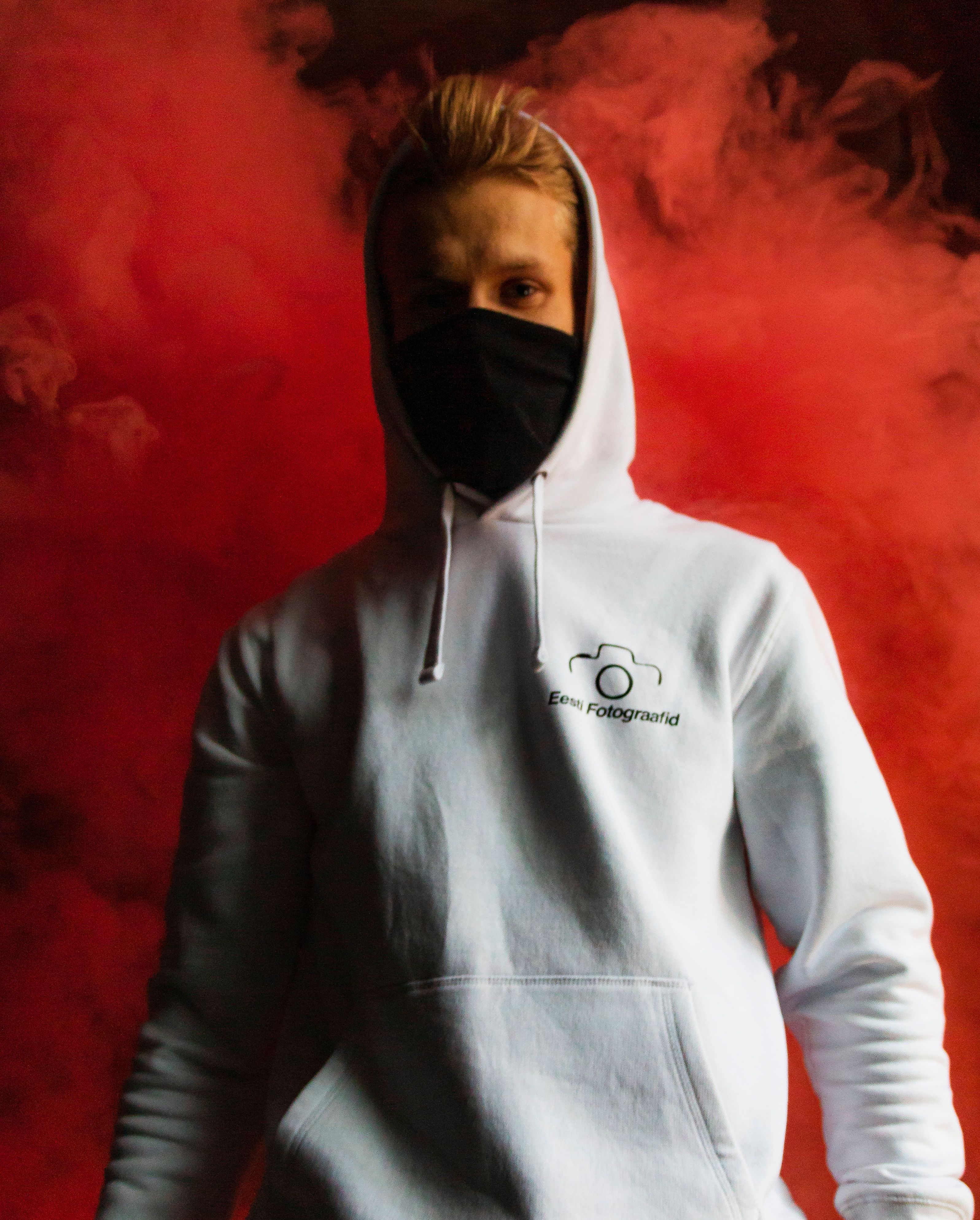 A person wears a face mask and hoodie against a red smoke backdrop, creating a dramatic portrait.