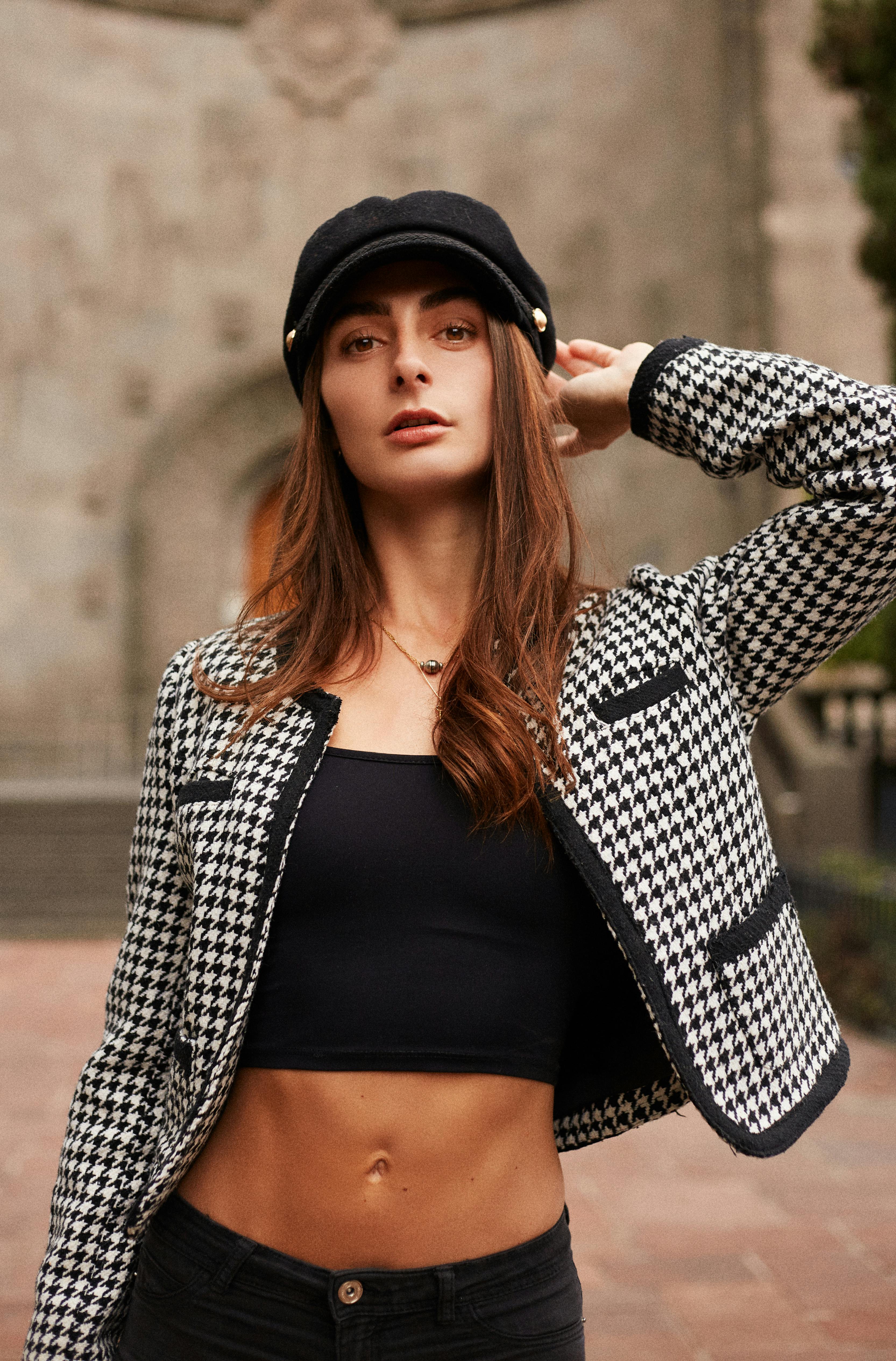 Stylish woman wearing a houndstooth jacket and cap, posing confidently outdoors.