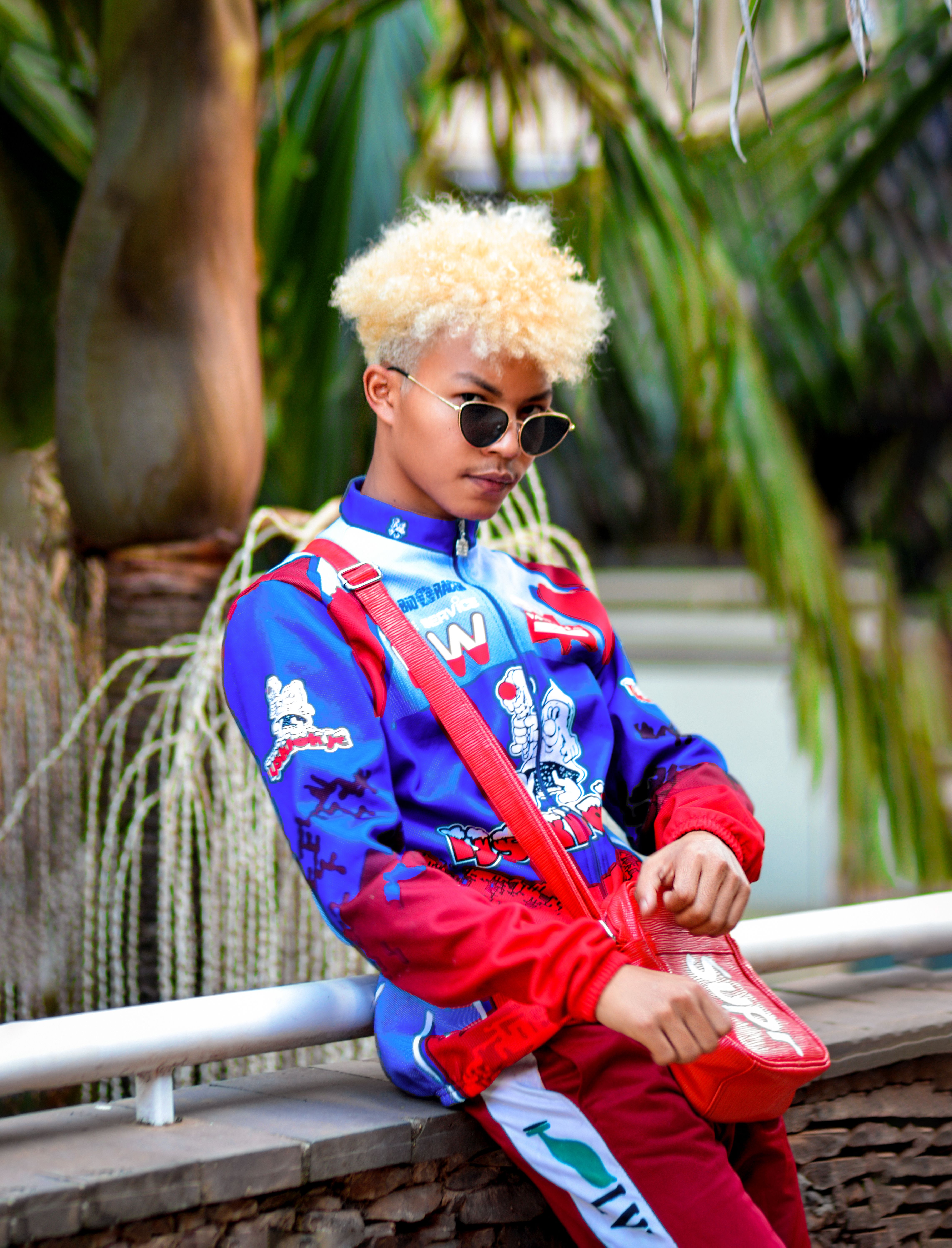 Stylish young adult in a colorful urban fashion leaning casually against a wall with lush greenery.