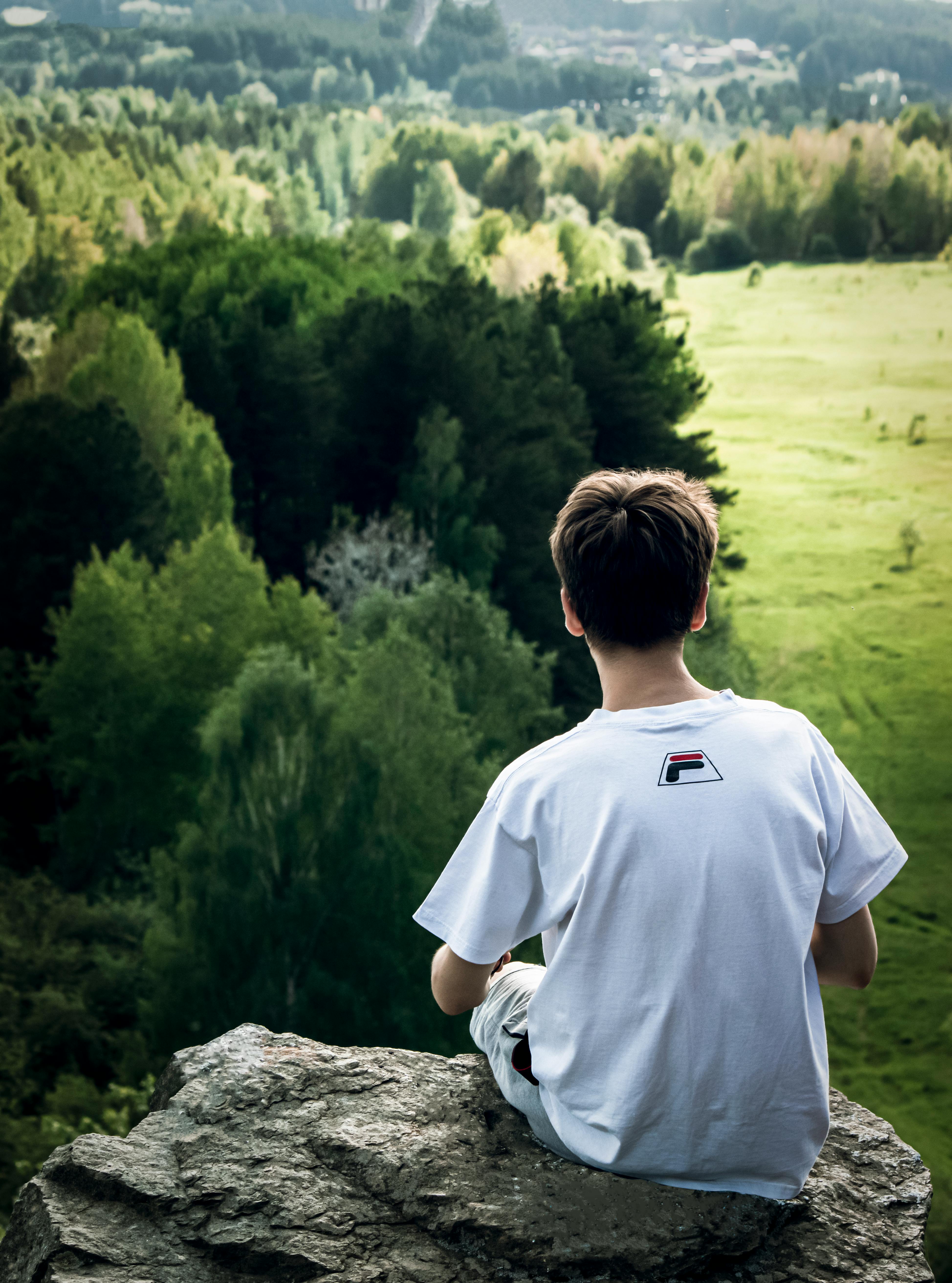 Young man sitting on a cliff, admiring a lush green landscape with vibrant forest below.