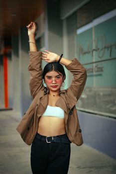 A confident young woman in a brown jacket posing with raised arms outside, showcasing fashion and style.
