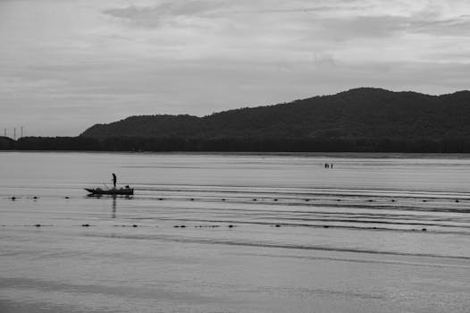 A fisherman in a boat on tranquil waters with distant mountains in Thailand.