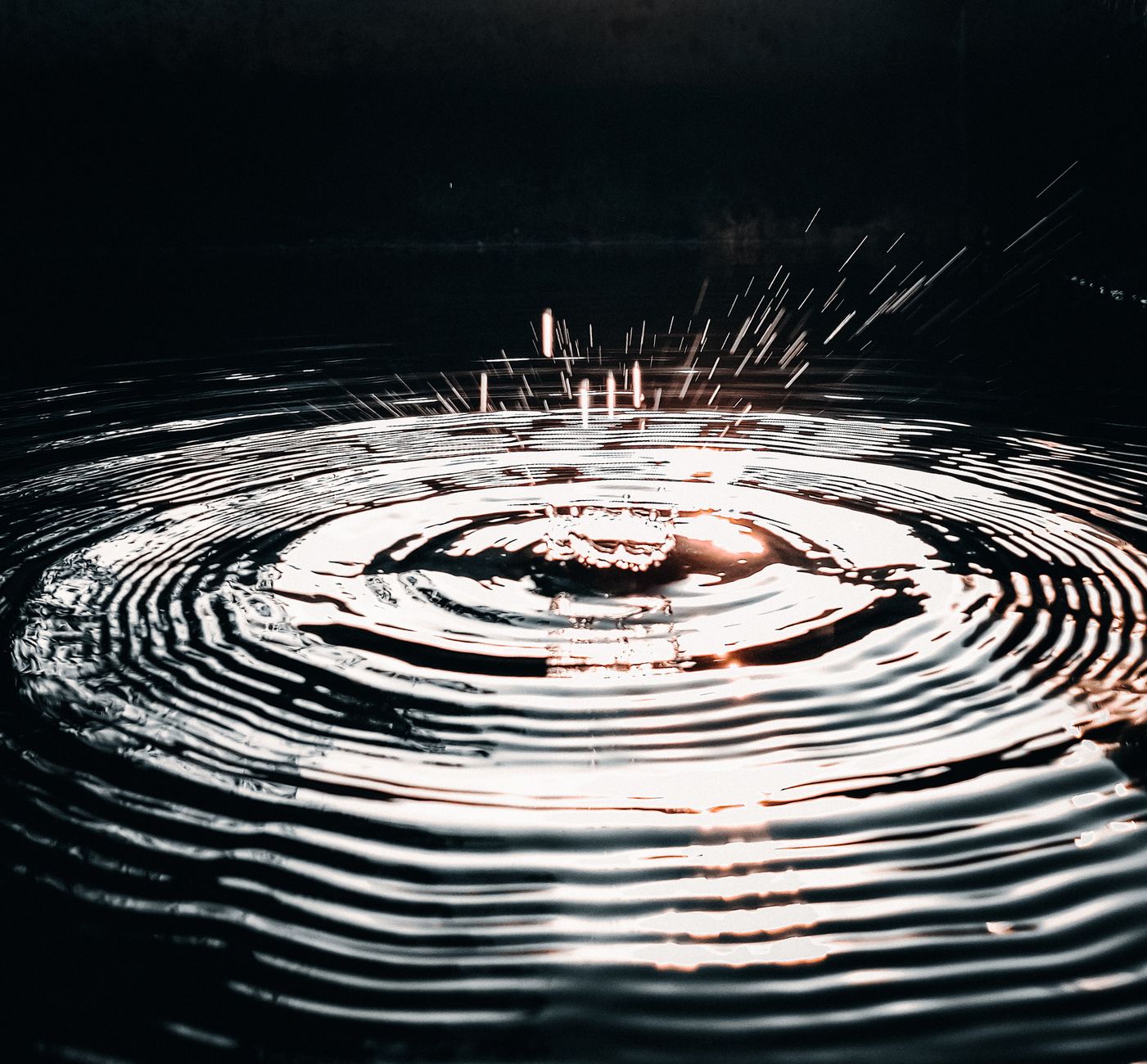 Detailed close-up of a water droplet causing ripples and splashes on a dark water surface, captured with striking light.