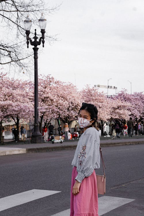 Young Woman Wearing a Face Mask Standing on a Street with Blossoming Cherry Trees in the Background