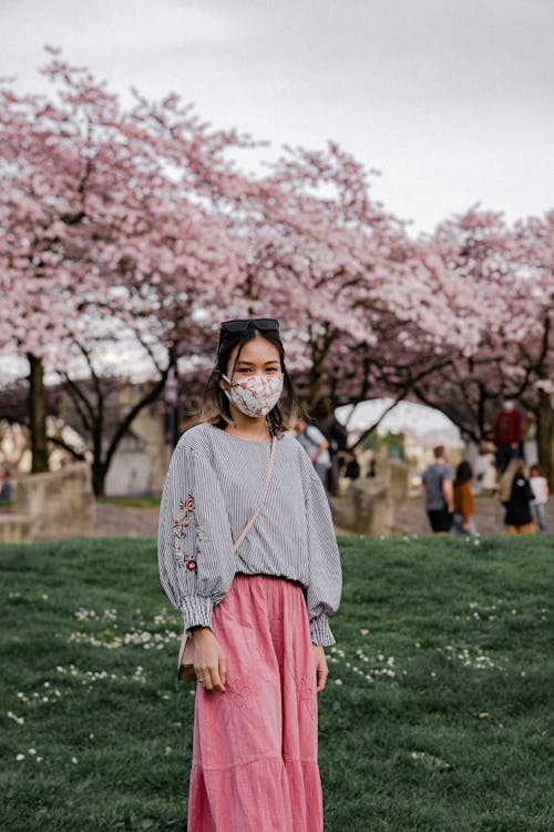 Young Woman Wearing a Face Mask Standing in a Park with Blossoming Cherry Trees