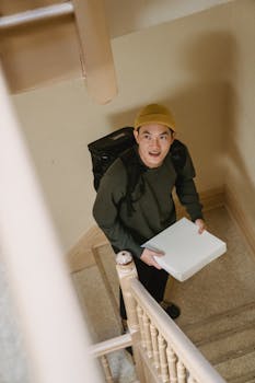 A young Asian deliveryman carrying a white box inside a building, looking upwards.