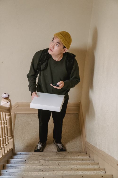 Free Courier in a cap holding a box and cellphone, standing on a staircase indoors. Stock Photo