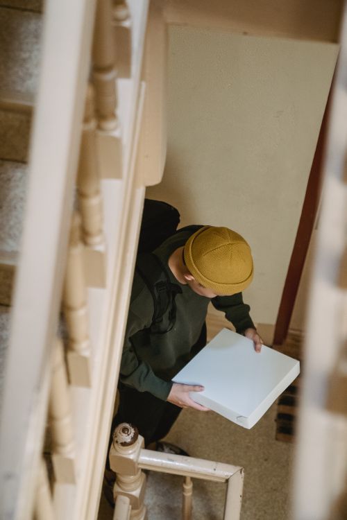 Free A delivery person in a beanie holding a pizza box indoors on a staircase. Stock Photo