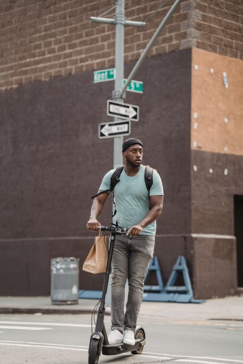 Free Young man with beanie riding an electric scooter on a city street, carrying a shopping bag. Stock Photo