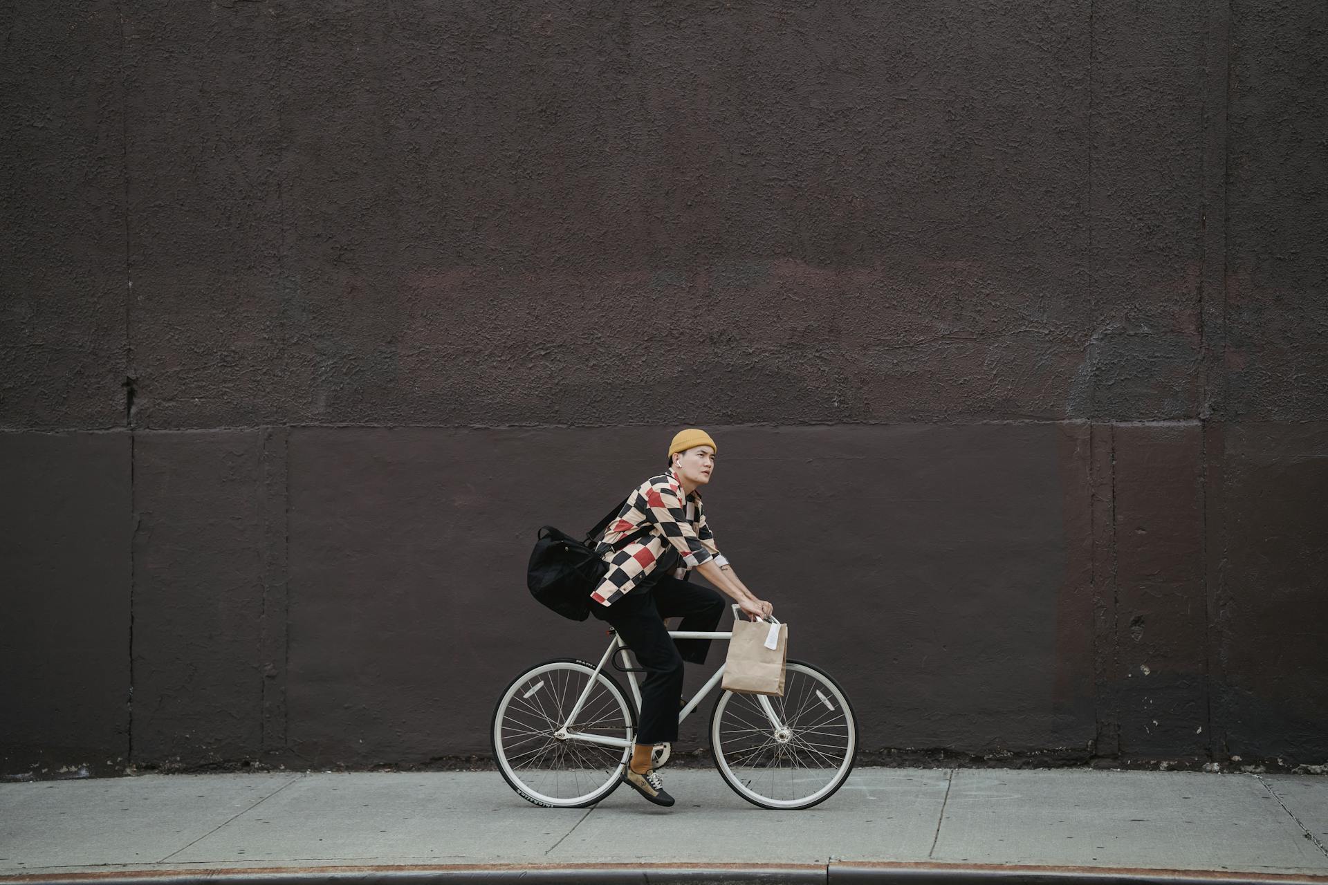 A delivery person riding a bicycle on an urban street against a large dark wall.