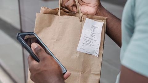 Man holding a paper shopping bag with a receipt, using a smartphone outdoors.