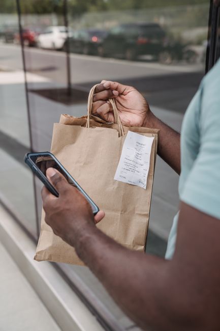 Man holding a paper shopping bag with a receipt, using a smartphone outdoors.
