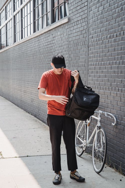 Man in Orange Crew Neck T-shirt and Brown Shorts Standing Beside Bicycle