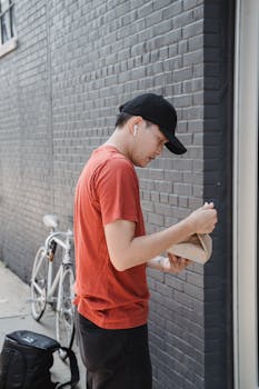 Asian man in casual attire, delivering a paper bag outdoors beside a brick wall.