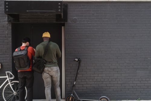 Free Two delivery men with backpacks at a brick wall entrance next to a scooter. Stock Photo