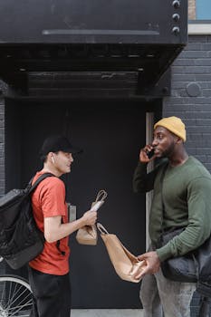 Two men exchange delivery bags at a building entrance, one on the phone.