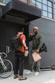 Two men with backpacks exchanging packages outside a building, one talking on a mobile phone.