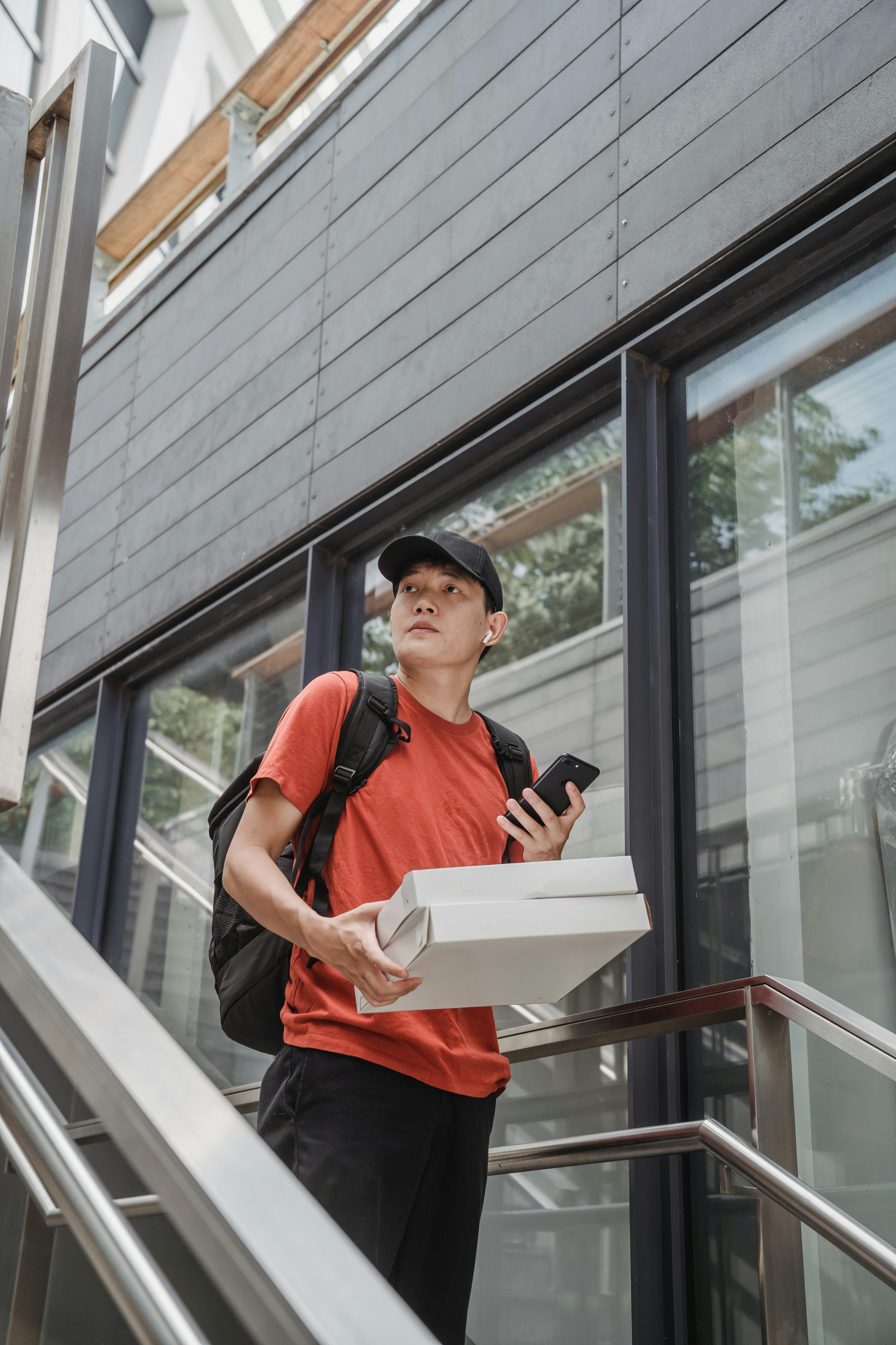 A pizza delivery person holds boxes and phone on a modern outdoor staircase.