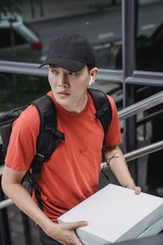 A delivery man with a backpack and cap carries a white box while looking aside.