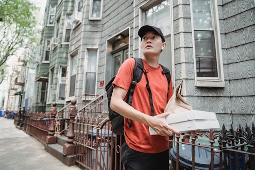 A delivery worker in a red shirt and cap holding packages outside a residential building.
