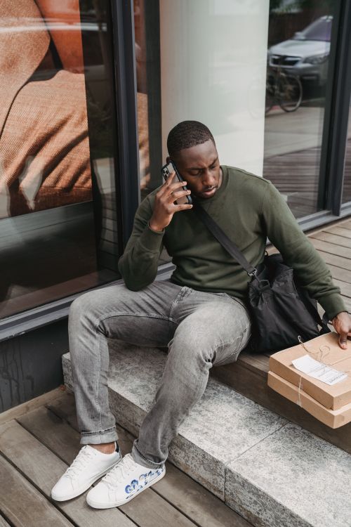 Free African American man talking on phone while sitting with delivery boxes outdoors. Stock Photo