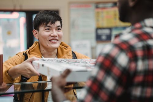 Free A cheerful Asian man receives a pizza box inside a shop, engaging with the seller. Stock Photo
