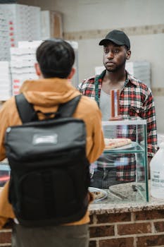 A customer with a backpack engages a vendor at a bakery counter indoors, showcasing a casual interaction.