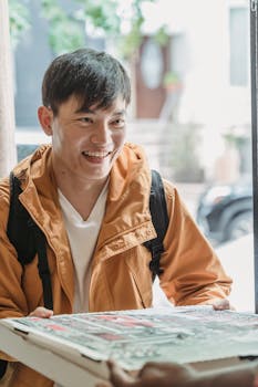 A cheerful delivery man in a jacket hands a pizza box to a customer, capturing a joyful pizza delivery moment.