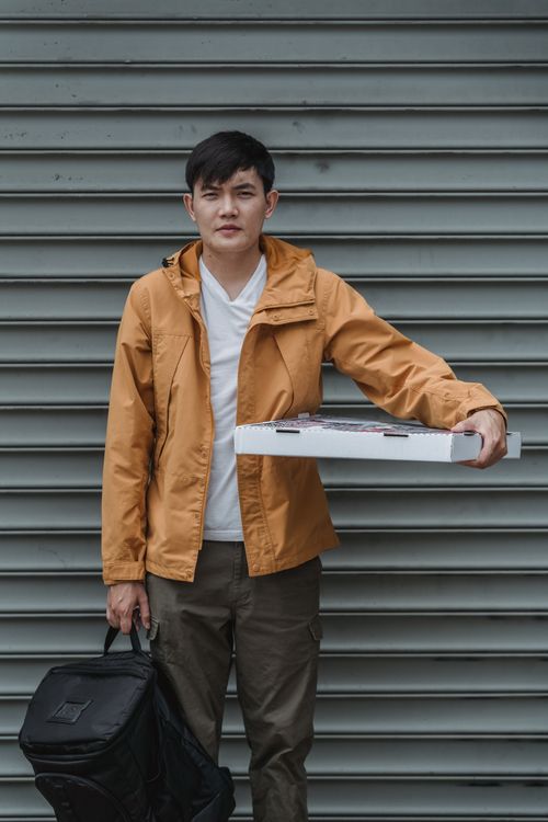 Free A delivery person in a hoodie holds a pizza box and thermal bag against a roller shutter background. Stock Photo